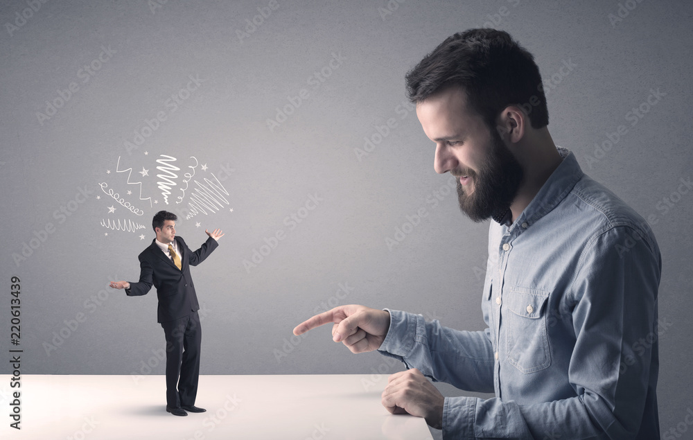 Young professional businessman being angry with an other miniature businessman who has scribbles above his head Young professional businessman being angry with an other miniature businessman who has scribbles above his head