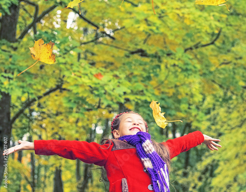 Little laughing pretty girl in red coat throws yellow leaves in autumn park