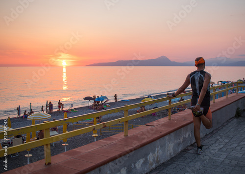 Uomo fa streching di frotne alla spiaggia al tramonto. Cielo arancione. Persone sulla spiaggia. Mare turchese. Montagna sullo sfondo