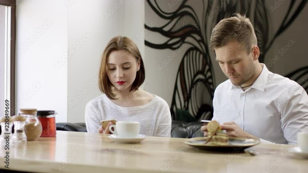 Two young people is sitting in a cafe. Man and woman are smiling and sitting in the cafe at the table during break. Couple is talking and drinking coffee. They are messaging on their mobile phones Two young people is sitting in a cafe. Man and woman are smiling and sitting in the cafe at the table during break. Couple is talking and drinking coffee. They are messaging on their mobile phones