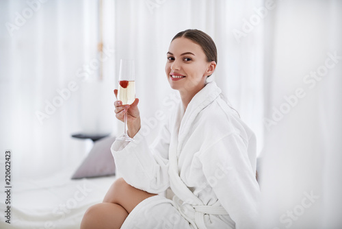 Taste this beverage. Smiling woman in bathrobe holding glass of champagne with strawberry
