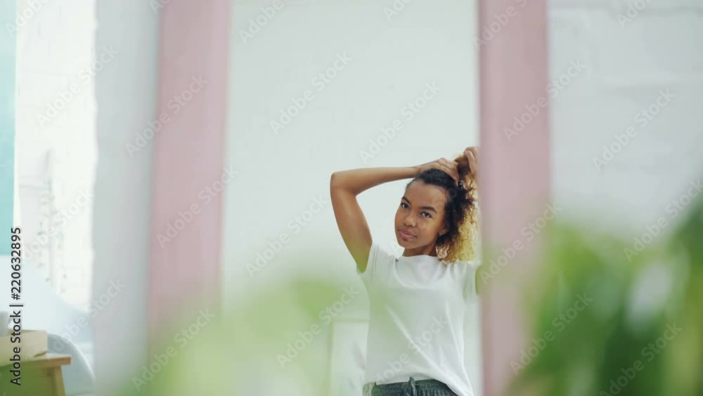 Pretty African American girl is fixing her curly hair looking in the mirror at home and smiling. Beautiful people, youth lifestyle and interiors concept. Pretty African American girl is fixing her curly hair looking in the mirror at home and smiling. Beautiful people, youth lifestyle and interiors concept.