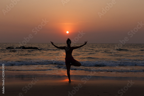 Young slender girl with her back doing yoga at sunset on the beach. Pose on balance, pose tree. Hands are raised and connected together. Natural backlight.