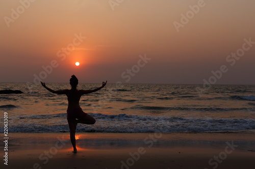 Young slender girl with her back doing yoga at sunset on the beach. Pose on balance  pose tree. Hands are raised and connected together. Natural backlight.