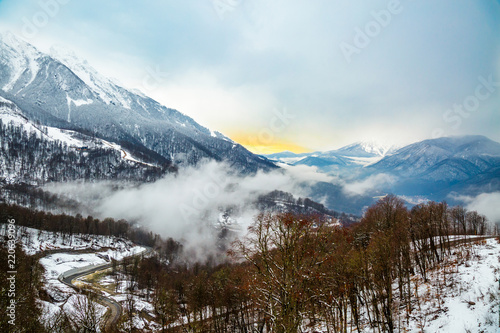 Foggy mountain landscape. Misty winter forest and valley. Fog at village, hiking famous place. View of snow capped mountains and cloudy sky at sunset in Rosa Khutor, Krasnaya Polyana, Sochi, Russia.