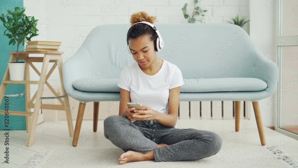 Pretty girl is playing game on smartphone touching screen and smiling while listening to music with headphones sitting on floor at home. Technology and fun concept. Pretty girl is playing game on smartphone touching screen and smiling while listening to music with headphones sitting on floor at home. Technology and fun concept.