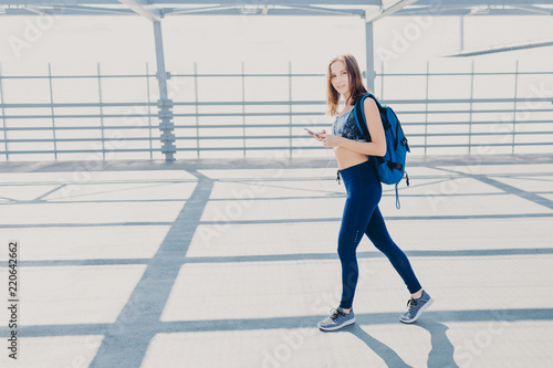 Pretty female youngster has walk outside, carries rucksack, holds modern cell phone, listens music, checks favourite song in playlist, wears casual sportswear and sneakers, has healthy lifestyle