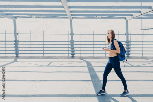 Outdoor shot of beautiful woman wth slim body, dressed in casual top and leggings, carries bag, going in gym, uses modern smart phone for listening music, copy space aside for your advertisement