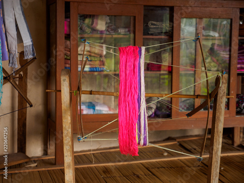 Close-up of dyed silk threads on a traditional spinning wheel with clothes and scarves in background Cabinets. Mae Chaem, Northern Chiang Mai, Thailand. Cottage industry and crafts.