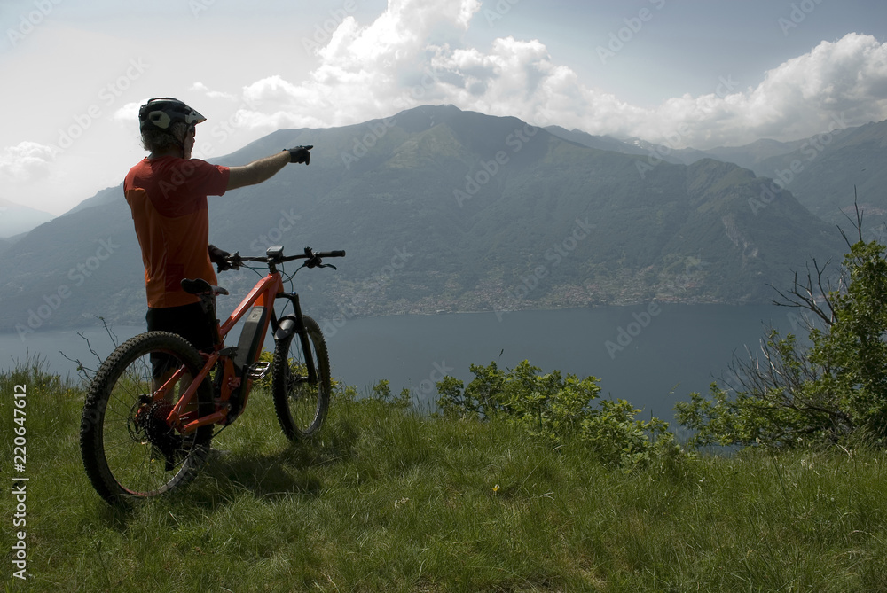 Fototapeta premium man with electric bicycle, e-bike, ebike, view of mountains, valley and lake of Como, summer, sport, adventure, cloud, sun, alps, Lombardy, Italy