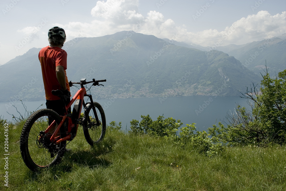 man with electric bicycle, e-bike, ebike, high mountains, observes horizon, valley, lake of Como, forest, summer, sport, adventure, cloud, sun, alps, Lombardy, technology, Italy