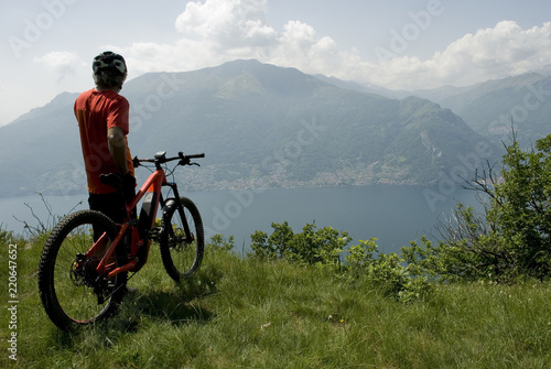 man with electric bicycle, e-bike, ebike, high mountains, observes horizon, valley, lake of Como, forest, summer, sport, adventure, cloud, sun, alps, Lombardy, technology, Italy