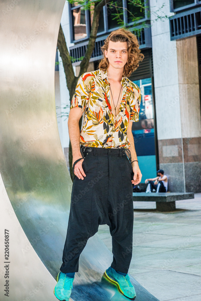 Portrait of Young American Artist with brown curly hair in New York,  wearing colorful patterned short sleeve shirt, baggy loose pants with  suspenders, patterned sneakers, hanging old key as necklace. Stock Photo