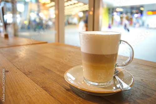 coffee latte macchiato in clear transparent glass and white plate lay on rough wooden table counter beside windows of coffee sop and blur background outside coffee shop.