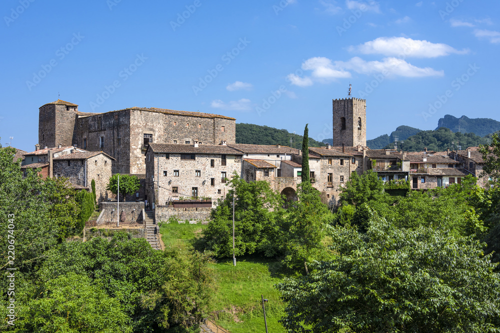Spain, Catalonia, Santa Pau: Panoramic view on the famous skyline of old ancient fortified Spanish town with tower, houses, green trees and blue sky in the background - concept travel medieval