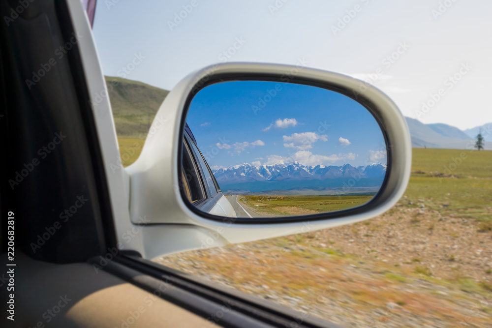 Reflection in the mirror of a riding white car of large altai mountains with snow on top by a sunny bright day with a blue clear sky green field and clouds