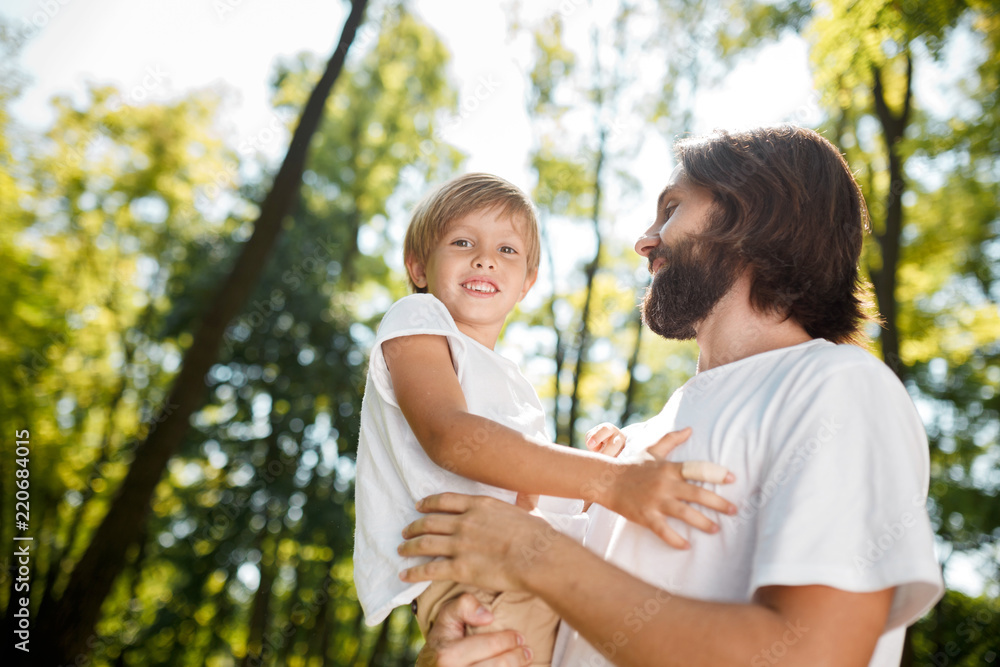 Handsome dark-haired father with beard dressed in the white t-shirt is holding in the arms his little son and watching at him on a summer day in the park. Handsome dark-haired father with beard dressed in the white t-shirt is holding in the arms his little son and watching at him on a summer day in the park.