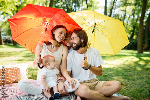 Cheerful parents with two kids have a rest on the lawn under the bright red and yellow umbrellas covering them from the sun.