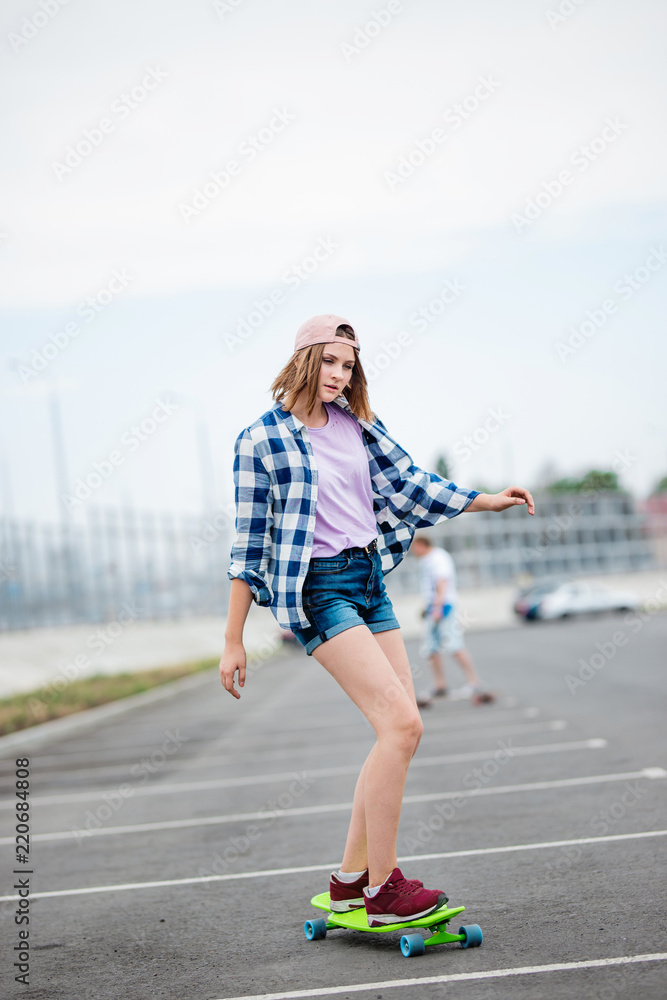 A beautiful blond girl wearing checkered shirt, cap and denim shorts is longboarding while stretching out her hands. Sport and cool style. A beautiful blond girl wearing checkered shirt, cap and denim shorts is longboarding while stretching out her hands. Sport and cool style.