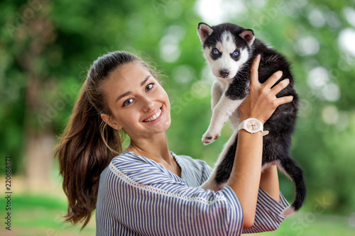 A beautiful smiling woman with a ponytail and wearing a striped shirt is cuddling with a sweet husky puppy while standing on the lawn. Love and care for pets.
