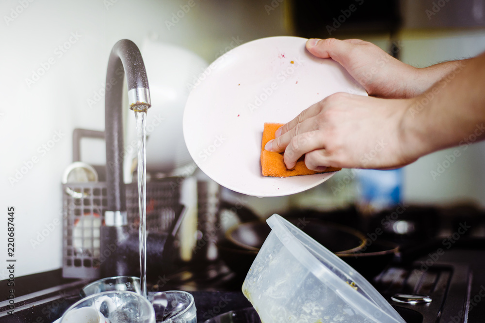The man washes the dirty dishes in the kitchen by hand. The man holds a dirty plate and a washing sponge in his hand. The concept of washing dirty dishes by hand. The man washes the dirty dishes in the kitchen by hand. The man holds a dirty plate and a washing sponge in his hand. The concept of washing dirty dishes by hand.