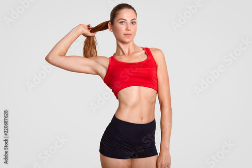 Horizontal shot of slim sporty woman has nice healthy muscular body, dressed in casual top and shorts, touches pony tail, poses against white background. Athletic girl takes break after workout