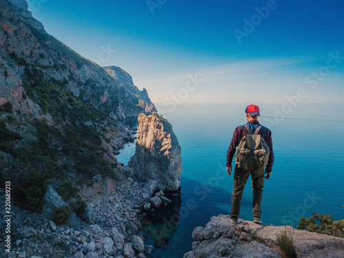 Young man backpacker with backpack standing on a rock cliff and enjoys views of the mountains and the sea. Hiker.