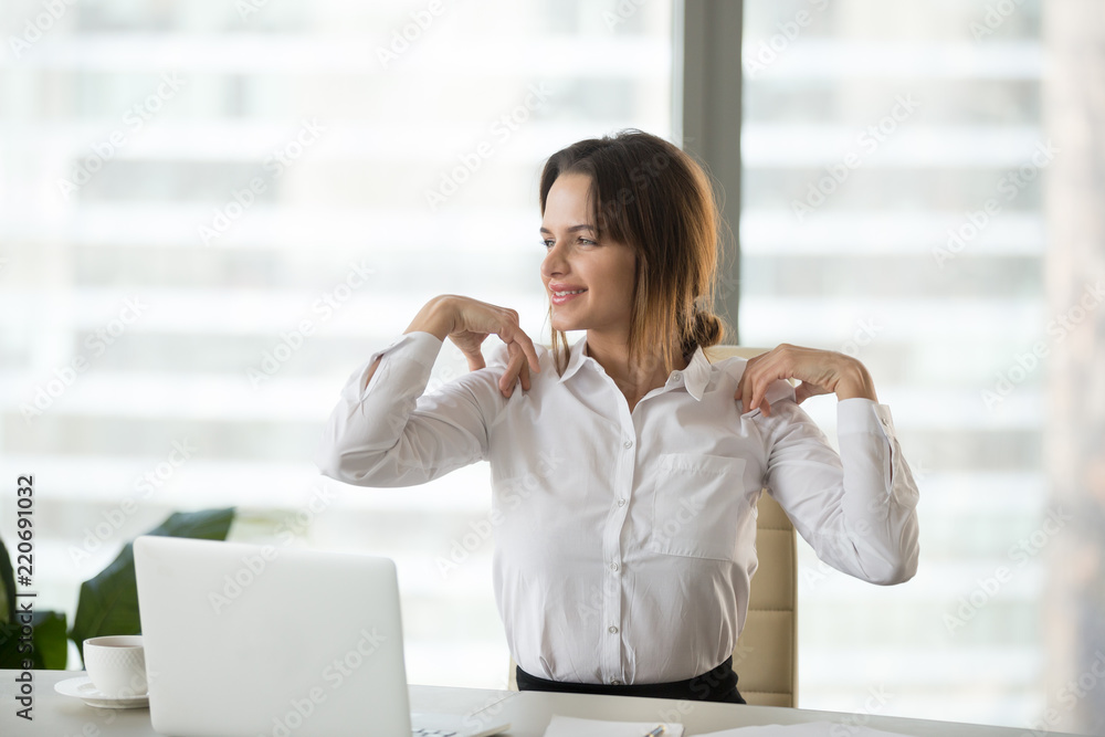 Smiling businesswoman doing easy office exercises to relieve neck and shoulder muscle tension from sedentary computer work, young employee taking break stretching for back relaxation at workplace Smiling businesswoman doing easy office exercises to relieve neck and shoulder muscle tension from sedentary computer work, young employee taking break stretching for back relaxation at workplace