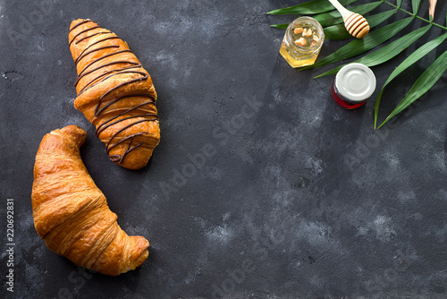freshly baked croissant decorated with chocolate sauce, jar with jams and palm leave isolated on a gray slate background. Top view with copy space