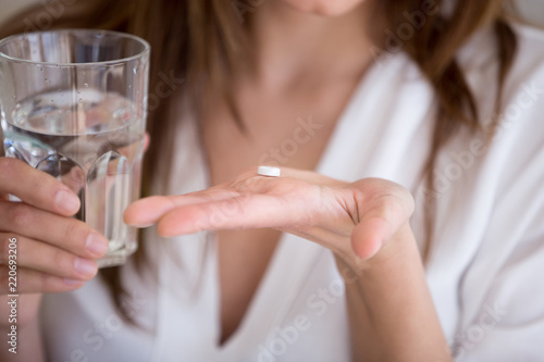 Woman holding pill and glass of water in hands taking emergency medicine, supplements or antibiotic antidepressant painkiller medication to relieve pain, meds side effects concept, close up view photo