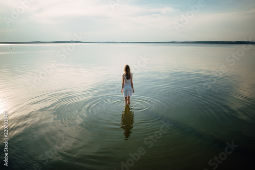 Woman in summer dress standing on seashore and looking at horizon. Young beautiful girl standing in water