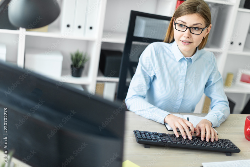 A young girl in glasses sits at a table in the office, holds a pencil in her hand and types the text on the keyboard. A young girl in glasses sits at a table in the office, holds a pencil in her hand and types the text on the keyboard.