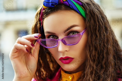 Outdoor close up fashion portrait of young beautiful woman wearing trendy violet sunglasses, colorful headband, posing in street
