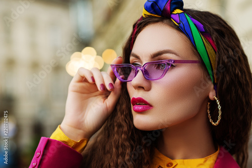 Outdoor close up fashion portrait of young beautiful woman wearing trendy violet sunglasses, colorful headband, circle earrings, posing in street of european city. Copy, empty space for text