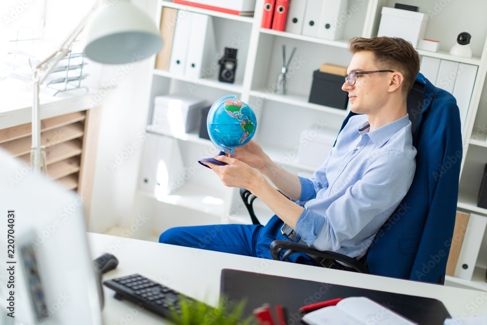 A young man sits in the office at a computer desk and holds a globe in his hands. A young man sits in the office at a computer desk and holds a globe in his hands.