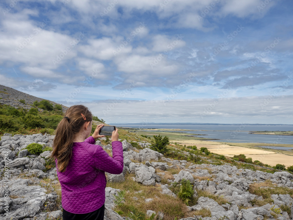 Young girl taking picture of beautiful landscape on her smart phone, Burren national park, Ireland. Concept travel memory, modern technology and use of smartphone during travel. Young girl taking picture of beautiful landscape on her smart phone, Burren national park, Ireland. Concept travel memory, modern technology and use of smartphone during travel.