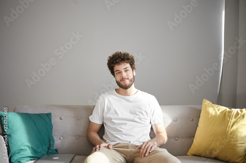 People, lifestyle, interior and design concept. Cheerful attractive young Caucasian man with stubble and stylish wavy hairdo sitting on comfortable couch with decorative pillows and smiling photo