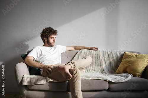 Horizontal shot of fashionable young thirty year old Caucasian bachelor with wavy hair and thick beard sitting comfortably on sofa in living room, having serious look, trying to relax after work photo