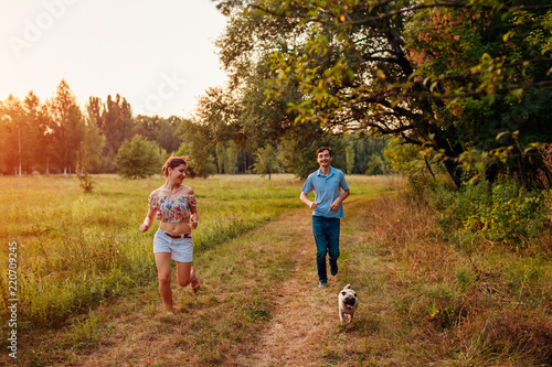 Young couple walking pug dog in autumn forest. Happy puppy running along and having fun playing with master.