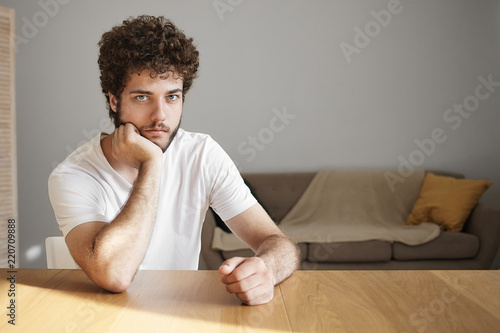 Horizontal shot of attractive serious young male with stubble having bored facial expression, spending weekend at home alone, sitting at wooden desk against white wall background, looking at camera