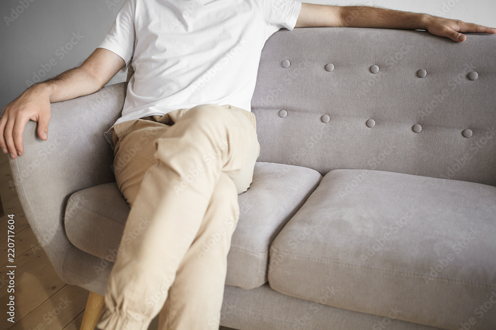 Cropped shot of unrecognizable man wearing white t-shirt and beige jeans sitting in the corner of comfortable cozy couch. Unknown male in casual clothes spending leisure time, relaxing on sofa