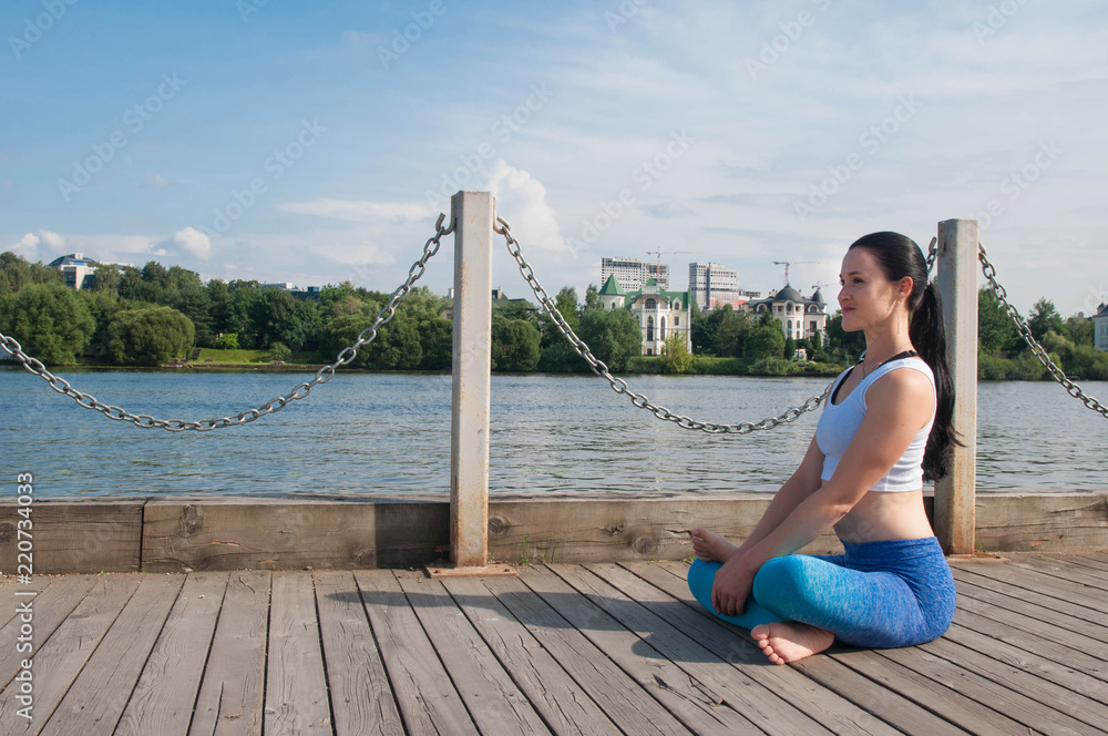 Young pretty fit and slim woman practices yoga and meditation on the embankment of the river