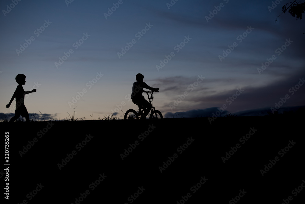 Silhouette of a boy riding a bicycle and another boy walking - outdoors