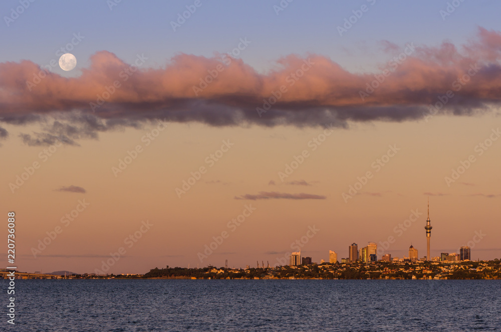 Full moon rising over the Auckland city skyline at sunset, looking across the water of the Waitemata harbour. Auckland, New Zealand.