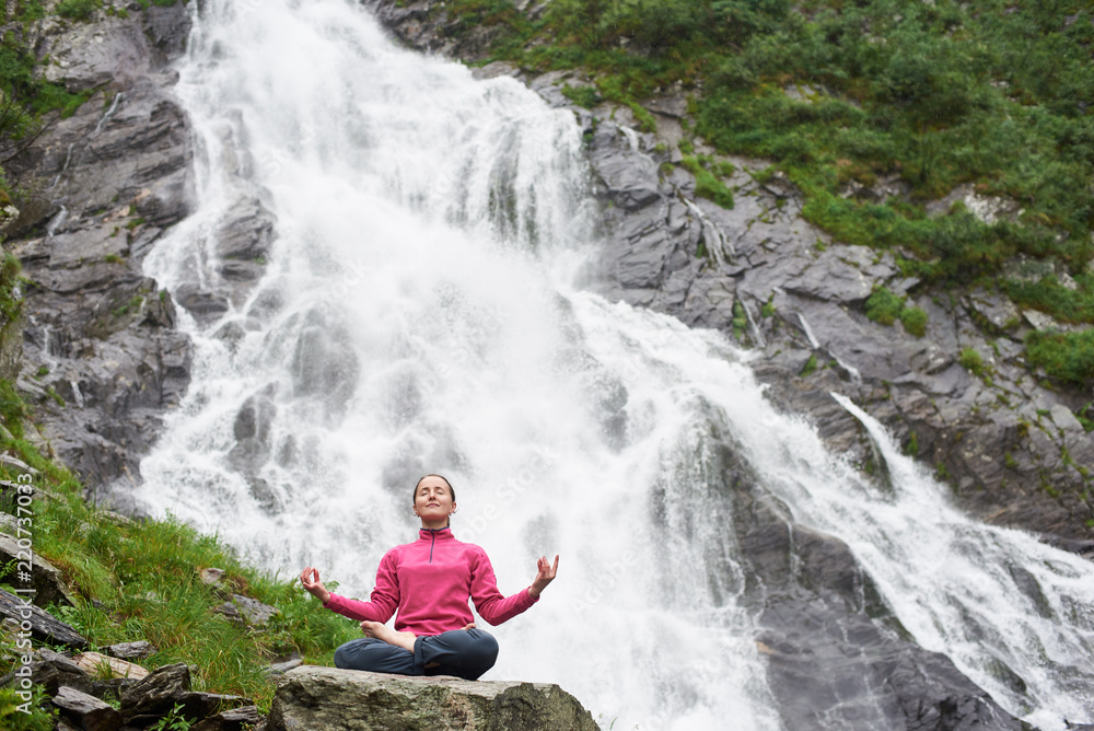 Young female practising yoga, meditating, relaxing alone against waterfall Balea in Fagarash mountains. Rocky waterfall in the mountains.