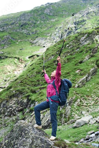 Close-up of tourist girl is standing with her arms wide open on a rock at the foot of the mountain. Girl with a backpack and trekking sticks. Fagaras Mountains, Carpathians, Romania