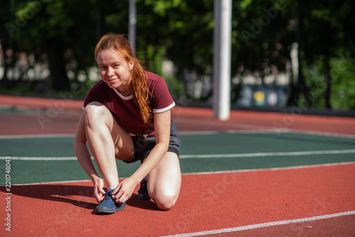 A young woman stopped to tie a string while running in the stadium. a red-haired student untied the lace during the lesson of physical education.
