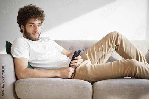 Portrait of handsome young European male with hairy face having rest on comfortable sofa, browsing newsfeed via social networks on his mobile phone, liking posts and leaving comments online