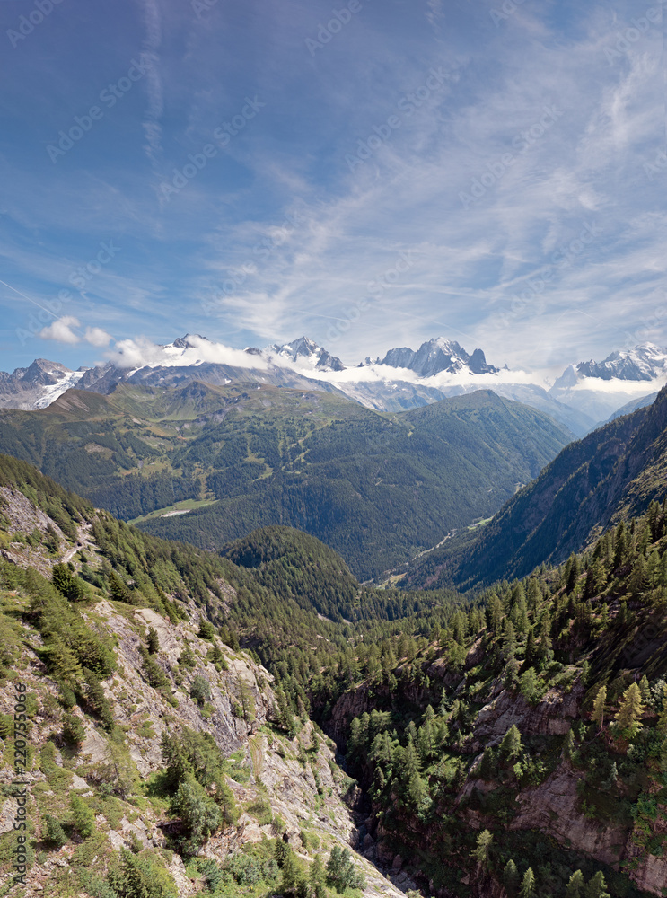Fototapeta premium Stunning snow capped peaks of the Alpine ridge of the Mont Blanc Massive. The village of chamonix is visible on the valley floor. The mountain sides are lush green with pine forests on a summer day.