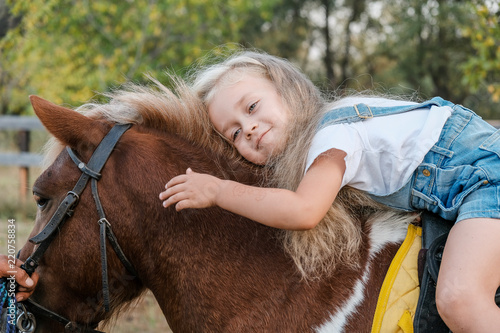 Fotografija A cute little blonde girl is sitting on a pony in autumn.